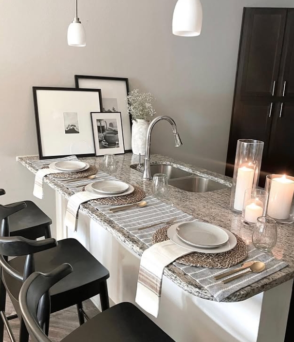 A modern kitchen island styled for a meal, featuring a granite countertop, white plates, glassware, woven placemats, and candles. Minimalist decor includes framed artwork and a bouquet, providing a warm and inviting atmosphere.