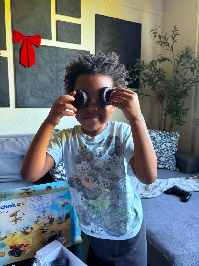 A young child playfully holding two cookie halves to their eyes, smiling and posing in a living room setting with a toy box and couch in the background.