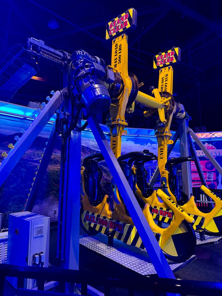 A close-up view of a yellow and black amusement park ride, featuring two ride seats, safety signage stating 'STAND CLEAR,' and mechanical components in a dimly lit environment with blue lighting.