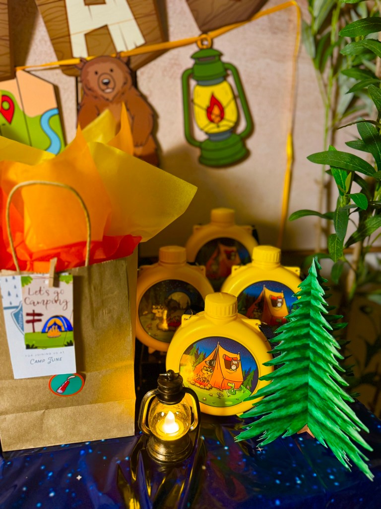 Camping party decorations with toy canteens, binoculars, paper pine trees, and cozy lantern lights.