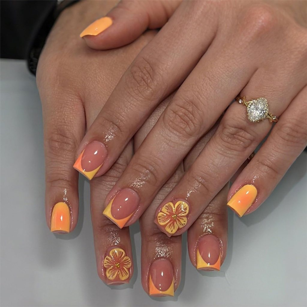 Close-up of two hands displaying nail art featuring orange and yellow geometric designs and floral accents, with a sparkling ring on one finger. The nails are glossy and manicured.