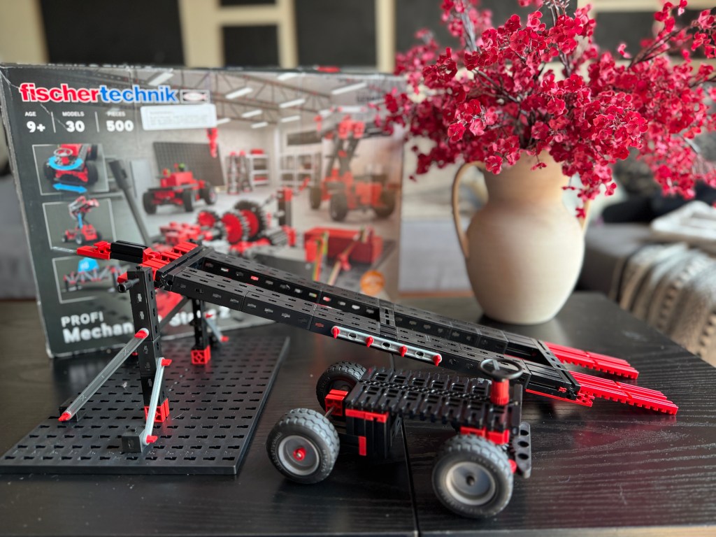A Fischer Technik STEM kit displayed on a black table, featuring a mechanical model with wheels and a conveyor belt design, alongside the kit's packaging and a vase of red flowers in the background.