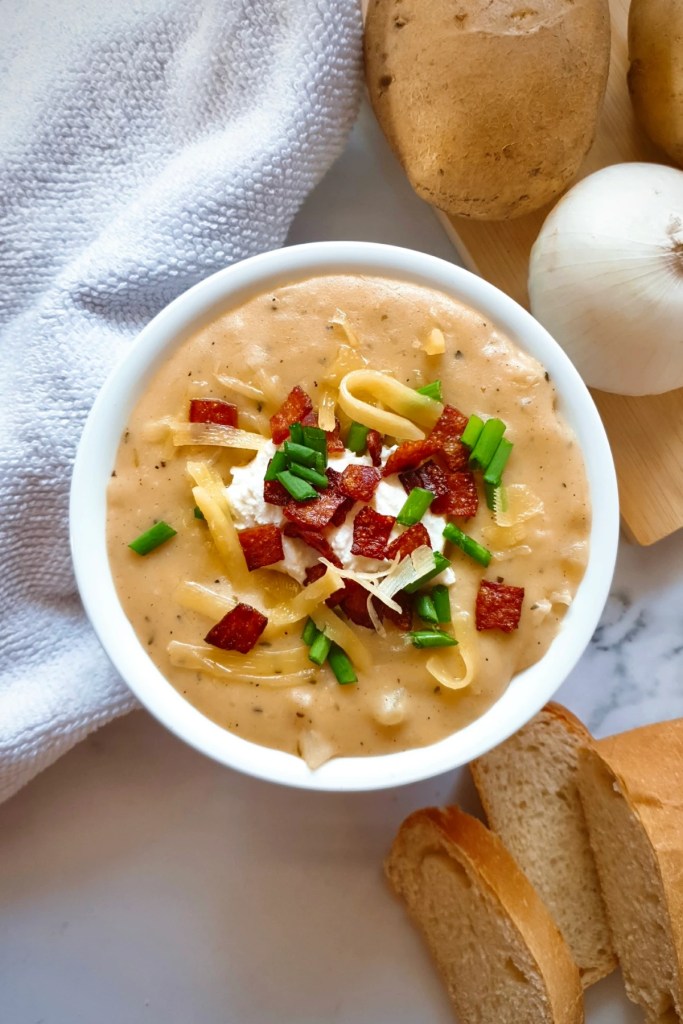 A warm bowl of dairy-free loaded baked potato soup topped with green onions, bacon bits, and creamy textures, served alongside fresh bread and potatoes.