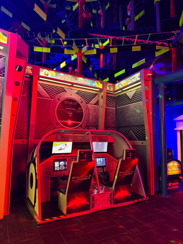 A brightly lit arcade area featuring a space-themed simulator with two cockpit-style seats and multiple screens displaying flight controls, surrounded by colorful neon lights.