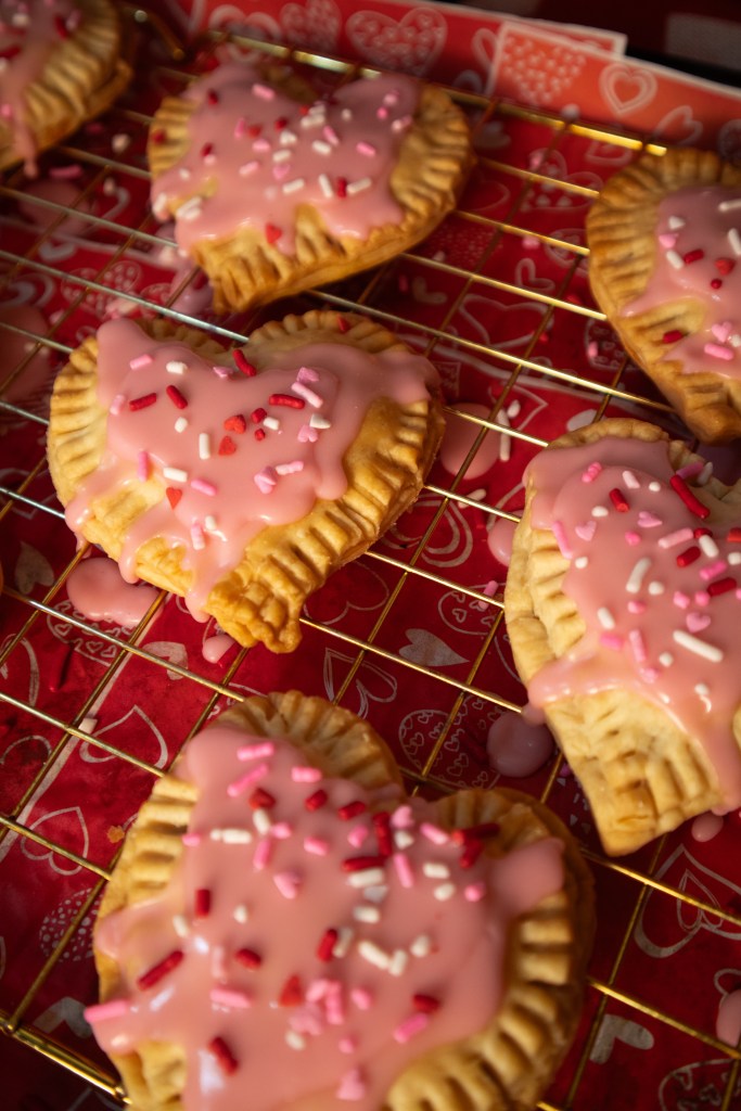 Baked heart-shaped pop tarts with pink icing and colorful sprinkles on a cooling rack, set against a decorative background.