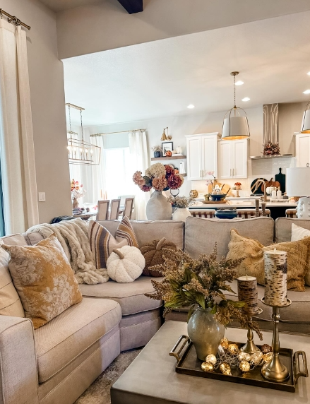 A cozy living room featuring a light-colored sectional sofa adorned with textured pillows and a warm throw. A coffee table showcases an arrangement of decorative items, including a floral centerpiece and candles, while soft lighting creates an inviting atmosphere.