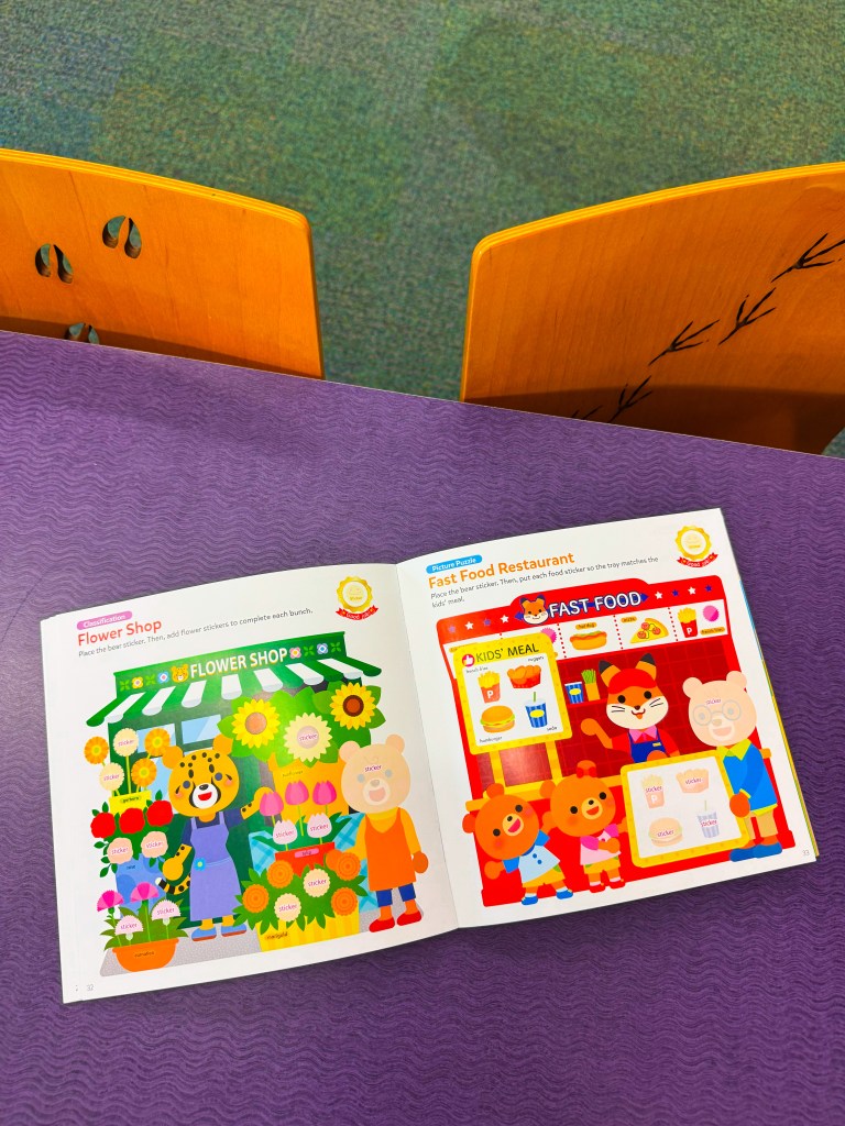 An open children's activity book displaying two pages: one featuring a colorful 'Flower Shop' illustration with flowers and cartoon characters, and the other page showing a 'Fast Food Restaurant' scene with food items and characters.