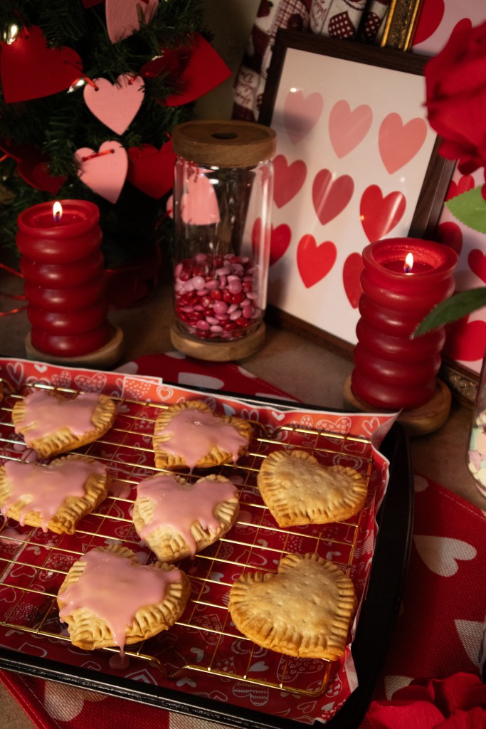 A tray of heart-shaped pop tarts with pink icing, placed on a decorative table adorned with red candles, a glass jar filled with colorful candies, and a picture frame featuring heart designs.
