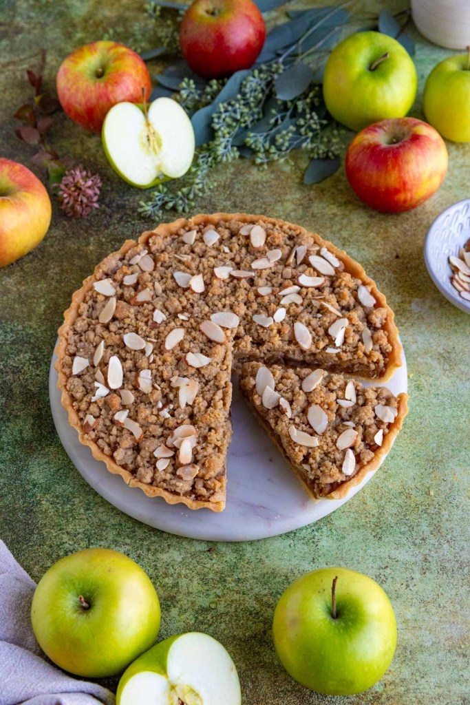 A sliced apple crumble tart garnished with almond slices on a marble plate, surrounded by red and green apples and greenery.