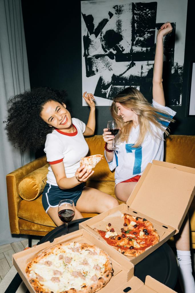Two friends enjoying pizza and wine while laughing and dancing on a cozy couch.