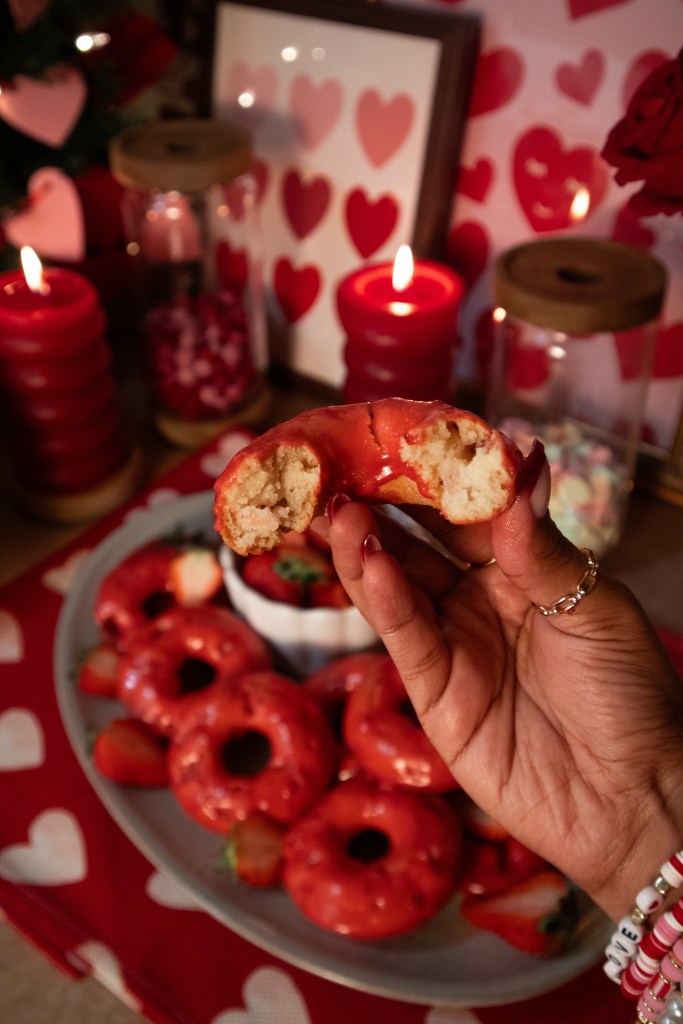A hand holding a half-eaten red strawberry glazed donut, surrounded by a plate of red donuts and strawberries, with decorative candles and heart-themed decor in the background.