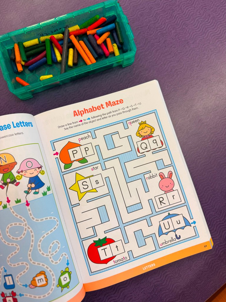 A colorful children's workbook page featuring an 'Alphabet Maze' with illustrations of various objects like a peach, star, rabbit, and umbrella. A container of crayons is positioned next to the book.