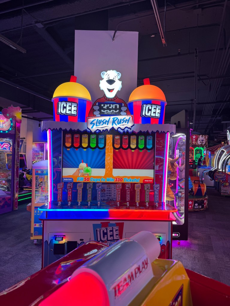 A brightly lit arcade game machine featuring the Slush Rush game, with colorful ice cream-themed design and various cup filling dispensers in an arcade setting.