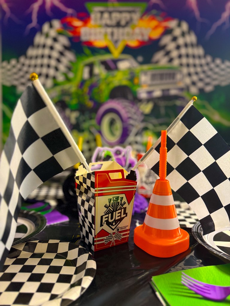 Racing themed monster truck birthday party table with checkered flags, traffic cone cup, tire plates, and party decorations.