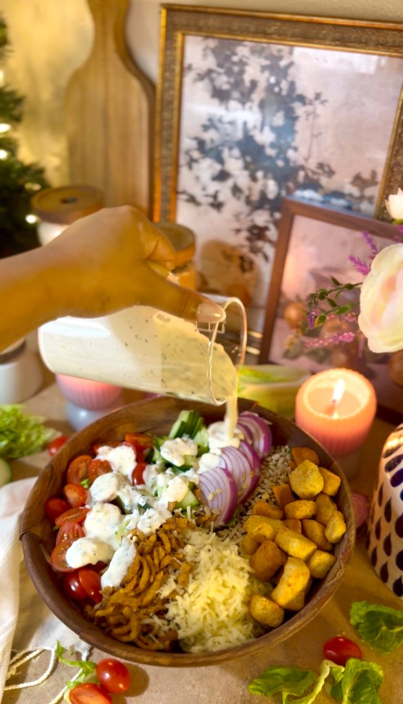 A hand pouring dressing from a glass container onto a colorful salad in a wooden bowl, featuring ingredients like cherry tomatoes, cucumber, red onion, rice, cheese, croutons, and garnished with herbs. In the background, lit candles and decorative elements create a cozy ambiance.