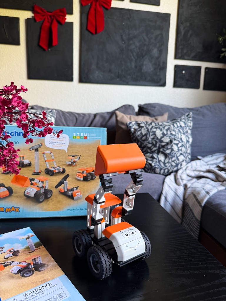 A colorful toy construction vehicle with a smiling face, positioned on a black table. In the background, a blue box for a construction set can be seen, along with a booklet featuring building instructions. The setting includes decorative red bows on the wall and a cozy sofa.