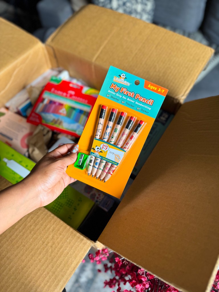 A hand holding a pack of colorful My First Pencil set designed for children aged 3-7, featuring thicker pencils for better grip, with a sharpener included, displayed in an open cardboard box filled with various school supplies.