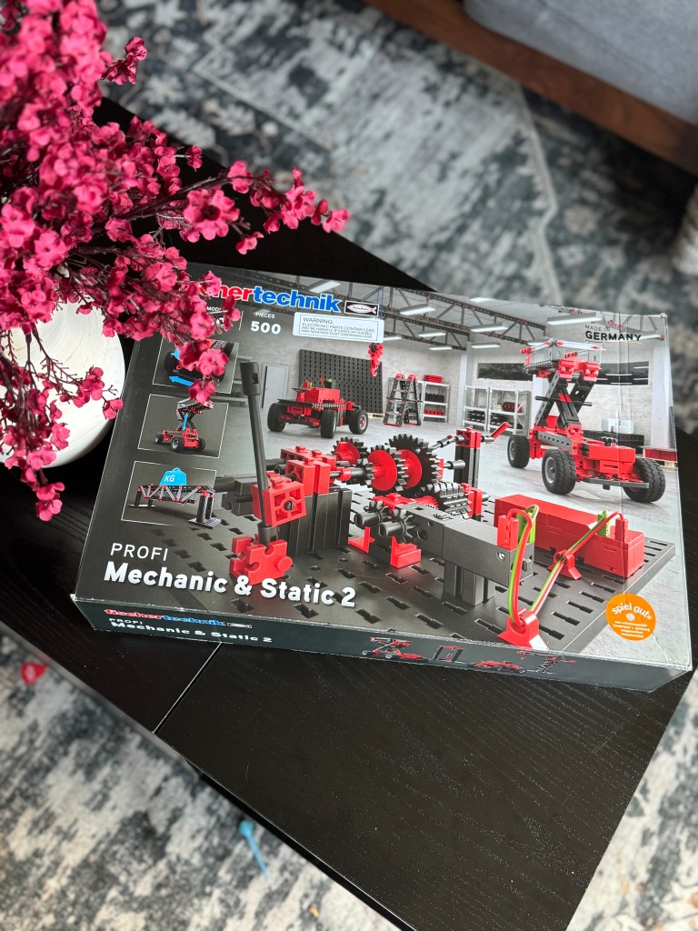 A box of the Fischer Technik STEM Kit featuring a design for mechanical and static building projects, with red and black components laid out on a table, surrounded by pink flowers.