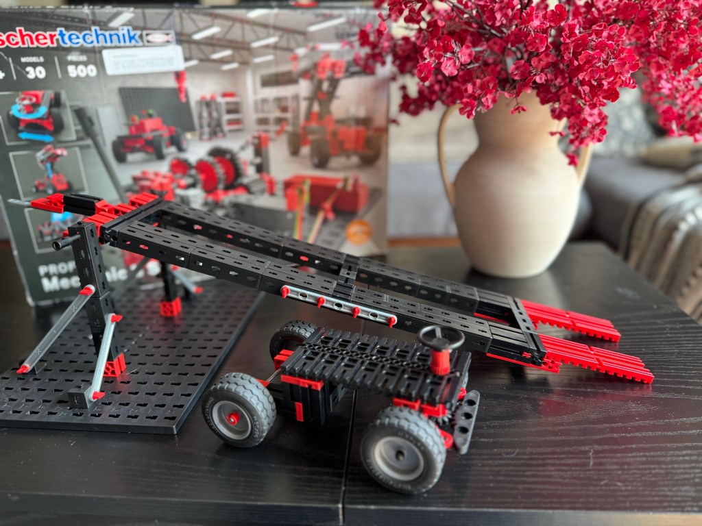A Fischer Technik STEM kit display featuring a built model of a vehicle with a ramp on a black table, surrounded by a decorative flower arrangement and a box showcasing other models.
