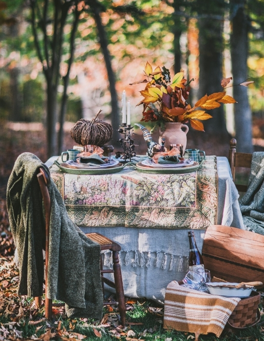 A beautifully styled outdoor Thanksgiving tablescape featuring a floral tablecloth, autumn leaves in a vase, and rustic dining elements like wooden chairs and a woven basket.