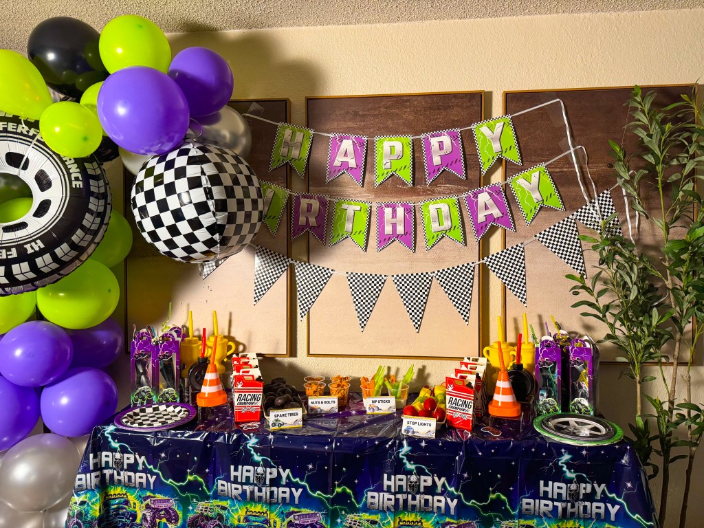 Monster truck birthday party setup with purple and green balloons, checkered racing decorations, and themed snack table for kids.