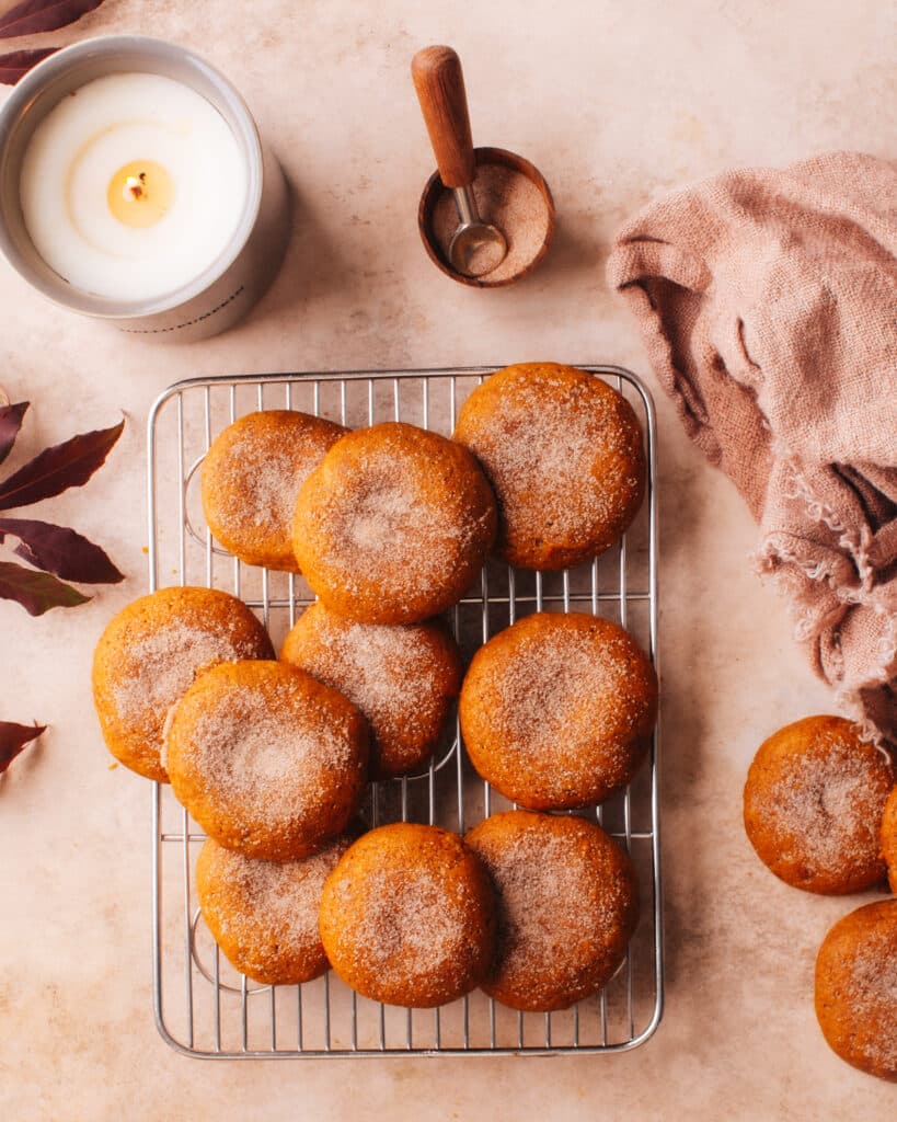 A cooling rack filled with freshly baked pumpkin cookies sprinkled with cinnamon sugar, surrounded by a candle, measuring spoon, and soft cloth.