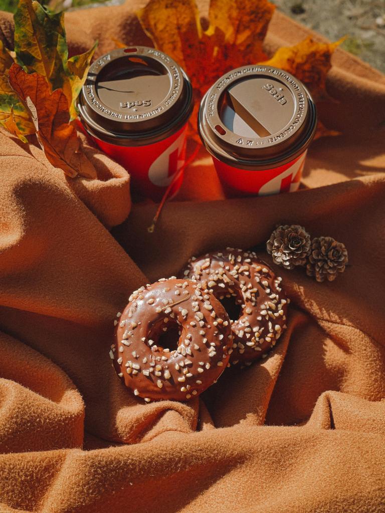 Two coffee cups with brown lids sitting on an orange blanket, surrounded by autumn leaves, two chocolate donuts topped with sprinkles, and a pine cone.