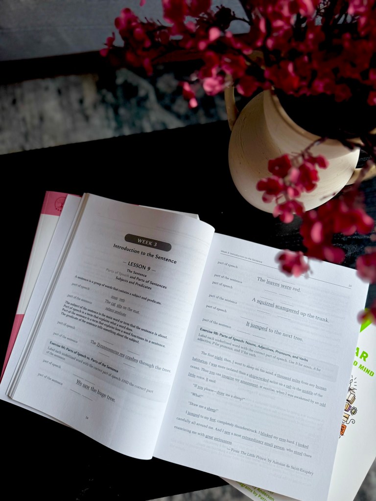 Open workbook page showing a lesson on sentence structure, featuring exercises about parts of speech, with a decorative vase of pink flowers in the background.