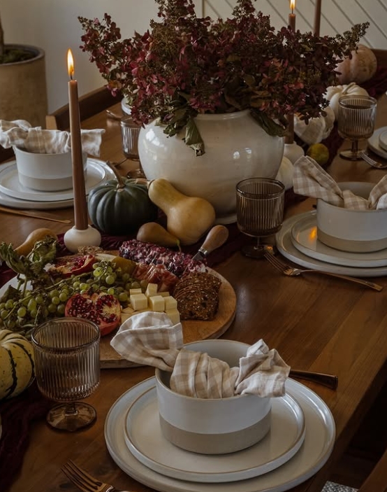 A beautifully arranged Thanksgiving table featuring natural textures and warm colors, including a centerpiece of dried flowers, candles, and styled dinnerware. The table is adorned with a variety of cheeses, fruits, and rustic elements, creating an inviting atmosphere.
