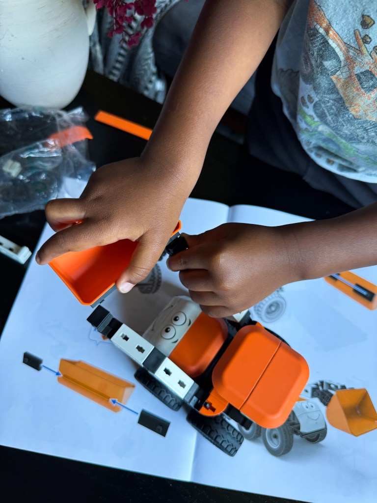 A child's hands assembling an orange toy truck while referring to build instructions on a booklet.