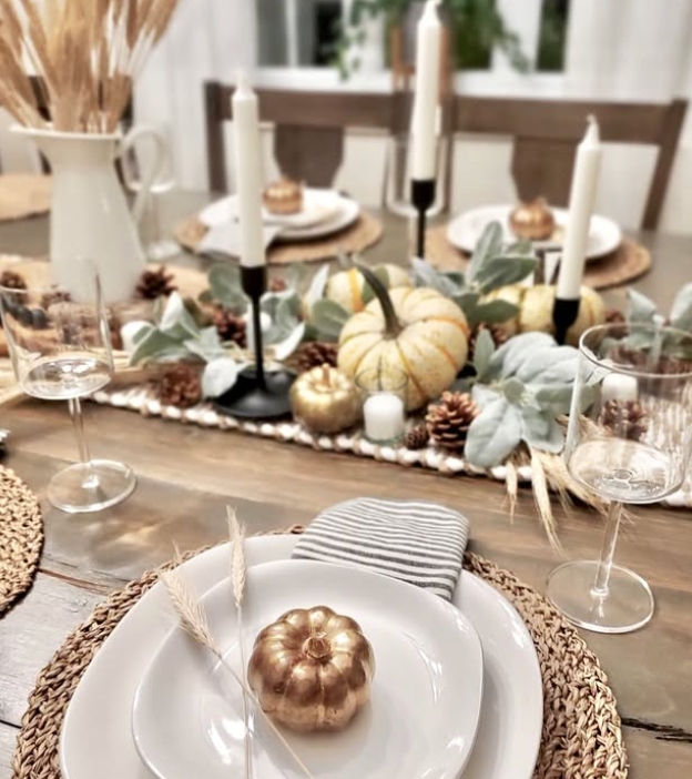 An elegant Thanksgiving tablescape featuring a wooden table topped with a decorative runner. The setting includes gold and white pumpkins, pinecones, and a decorative arrangement of greenery. White plates are surrounded by woven placemats, with candles in black holders providing ambient lighting.