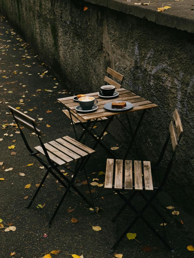 A small wooden table set for two, with a blue cup, a plate of cake, and a latte, surrounded by fallen autumn leaves on a cobblestone path.