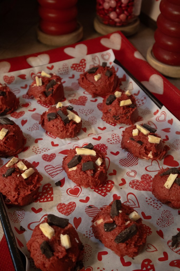 A tray of red velvet cookie dough balls topped with white and dark chocolate pieces, resting on heart-patterned parchment paper.