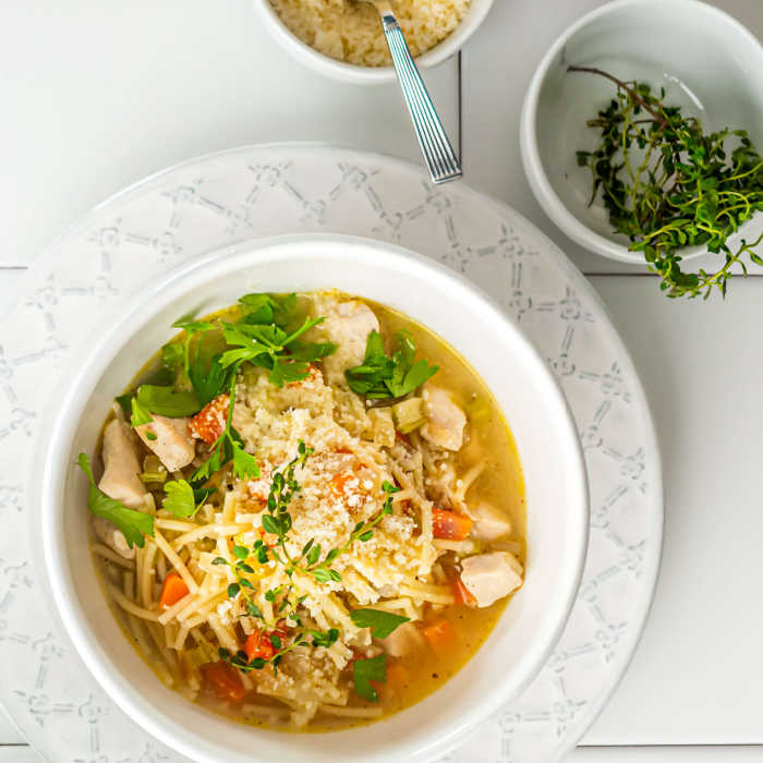 A warm bowl of chicken noodle soup topped with herbs and cheese, served on a decorative plate alongside bowls of grated cheese and fresh thyme.