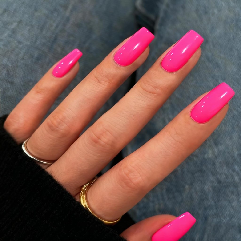 Close-up of a hand with long, bright pink nail polish on the nails, resting on a textured surface, showing a black sweater sleeve and multiple rings on the fingers.