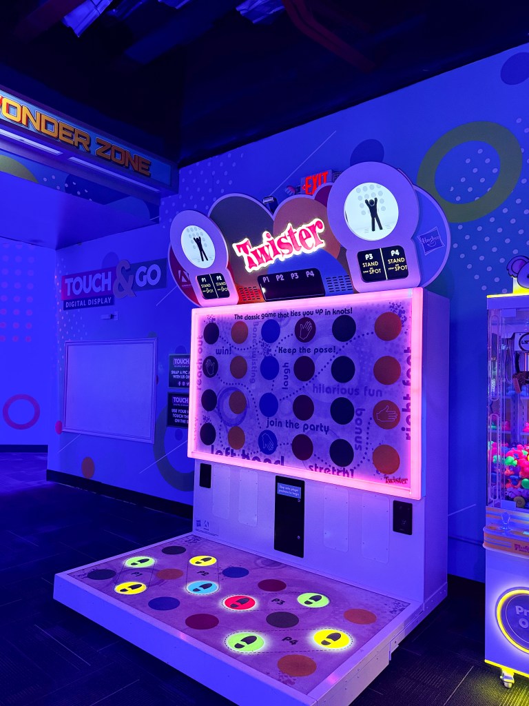 A digital Twister game display with colorful circles and instructions, set in a vibrant gaming area with purple lighting.