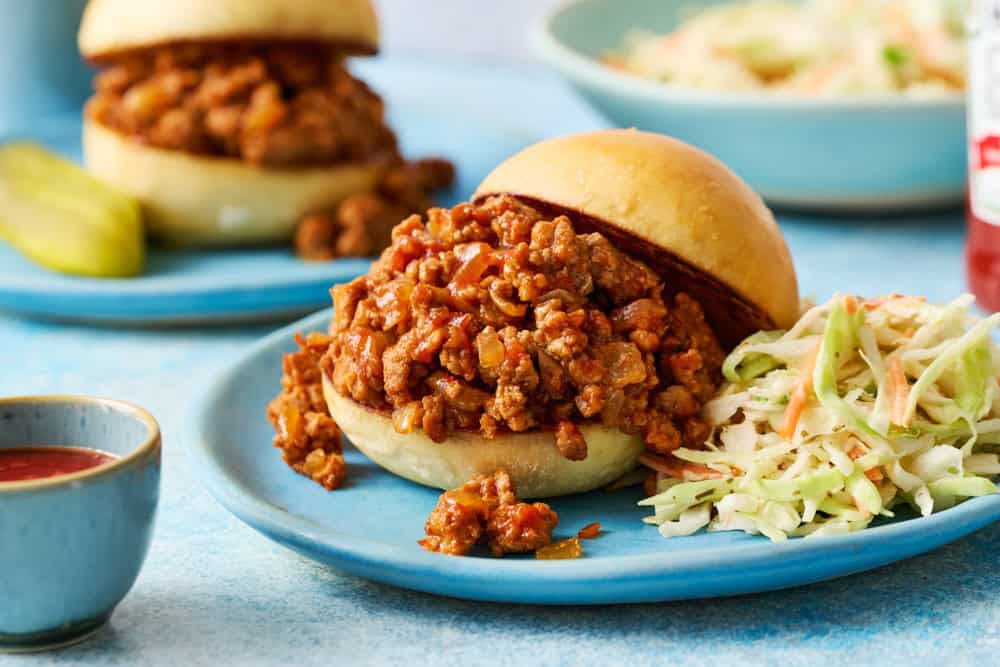 A close-up of a ground chicken sloppy joe sandwich on a blue plate, accompanied by a side of coleslaw and a small bowl of sauce.