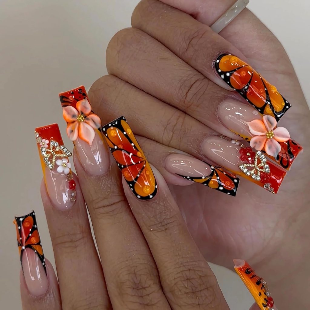 Long acrylic nails with orange polish, black detailing, and 3D floral accents