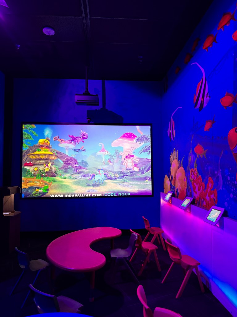 Colorful room featuring a large screen displaying a vibrant animated scene with fantastical creatures and mushrooms, surrounded by playful furniture including a pink table and colorful chairs.