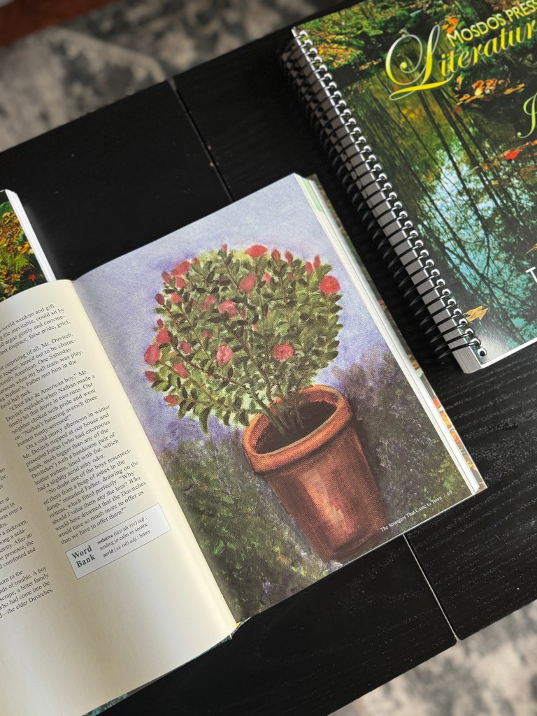 An open literature book featuring a colorful illustration of a flowering plant in a terracotta pot, placed on a wooden table next to a spiral-bound textbook.