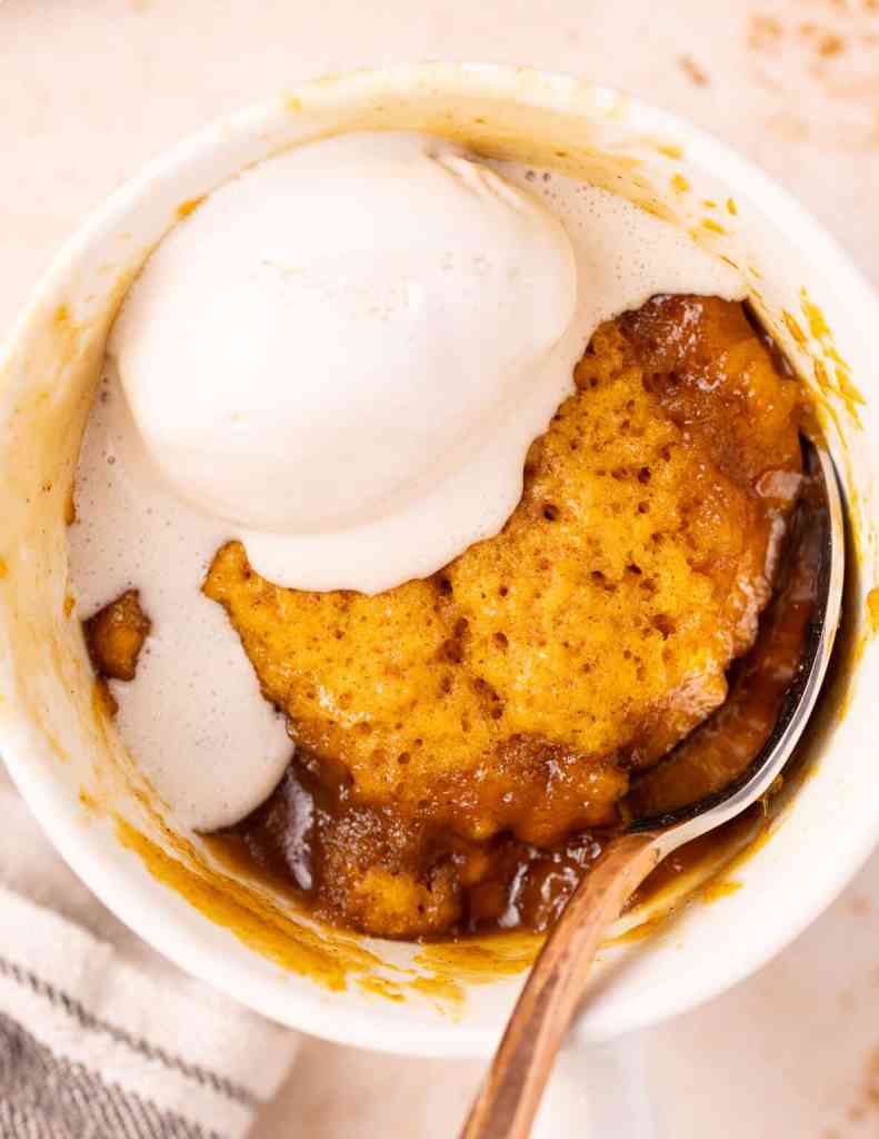 A warm pumpkin mug cake topped with a scoop of vanilla ice cream, served in a white mug with a metal spoon.