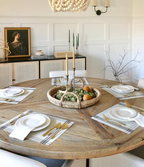A beautifully styled round dining table set for Thanksgiving with a woven centerpiece, candles, and elegant dinnerware. Gold utensils and plaid napkins create a warm and inviting atmosphere.