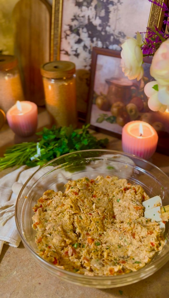 A glass bowl filled with a mixed dish for salmon patties, surrounded by candles, herbs, and decorative items, set on a table.