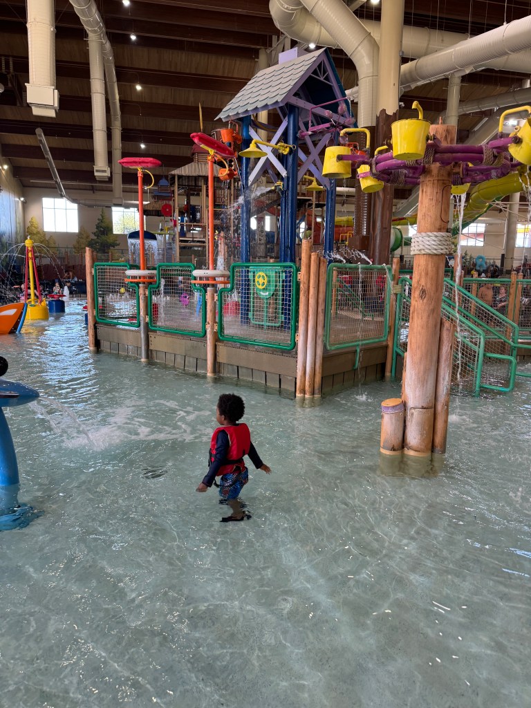 A child wearing a life jacket playing in a shallow area of an indoor water park, featuring colorful play equipment and water features.