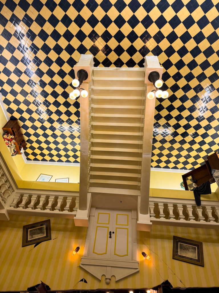 An upside-down staircase viewed from below, leading to a yellow door, with a checkered ceiling and decorative wall patterns.