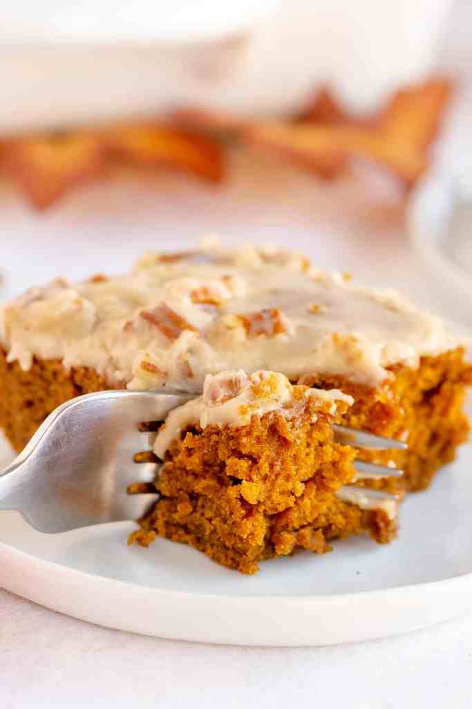A close-up of a moist pumpkin cake topped with cream cheese frosting, served on a white plate with a fork beside it.