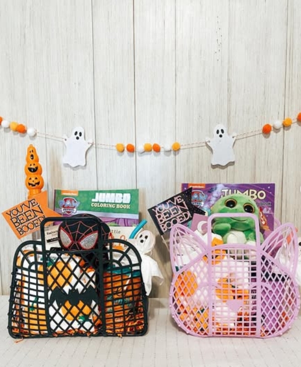 Two Halloween-themed boo baskets filled with treats and toys, one decorated with an orange and black spider web design and the other in pastel pink, placed against a white wooden backdrop with decorative ghosts overhead.