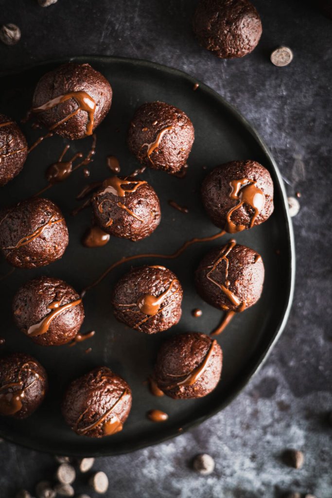 A close-up view of chocolate date balls drizzled with chocolate on a black plate, surrounded by chocolate chips.