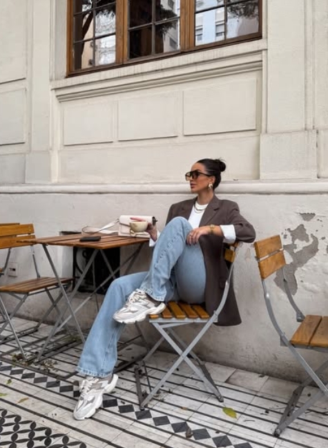 A woman seated at a café table, wearing a brown blazer over a white top and light blue jeans, accessorized with sunglasses and sneakers. She is sipping coffee, exuding a relaxed yet stylish vibe.