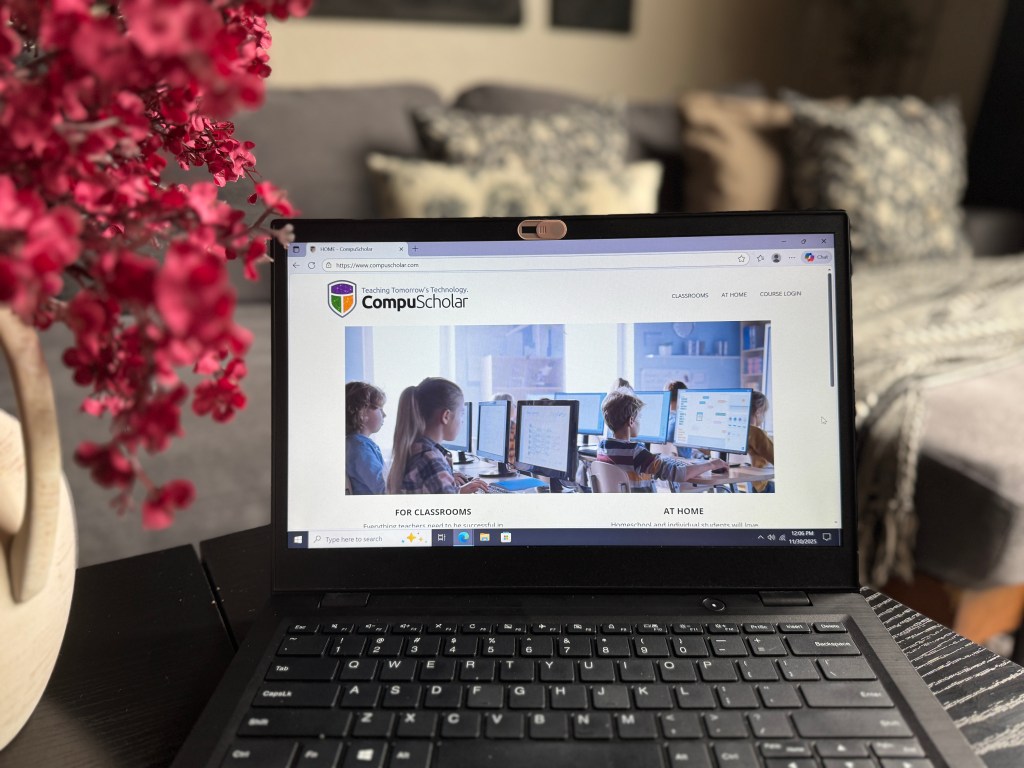 A laptop displaying the CompuScholar website, featuring students engaged in tech learning activities. A potted plant with pink flowers is in the foreground.